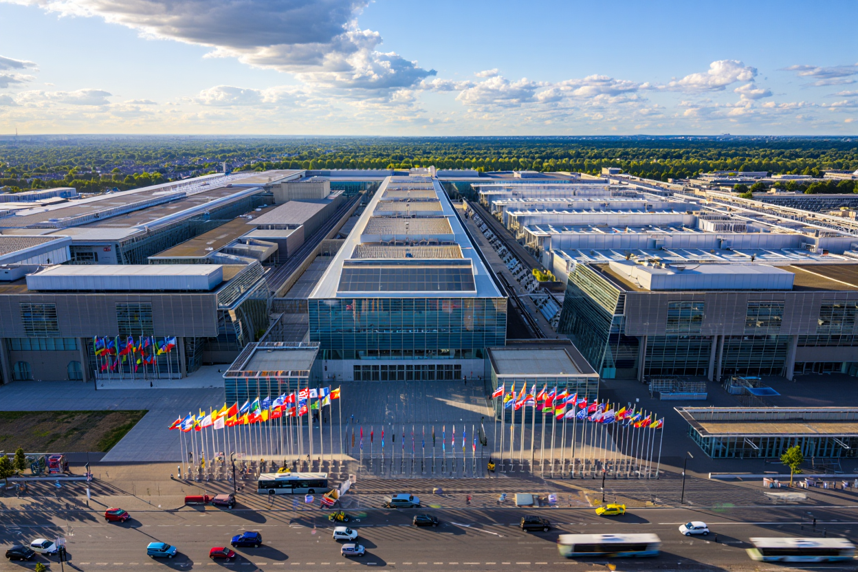 Grand ensemble de halls modernes vus d’en haut, façade vitrée et drapeaux multicolores devant l’entrée principale, avenue avec voitures. salon objet publicitaire paris?